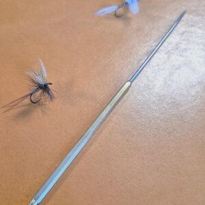 A fishing fly beside a thin, white fishing rod on a wooden surface.